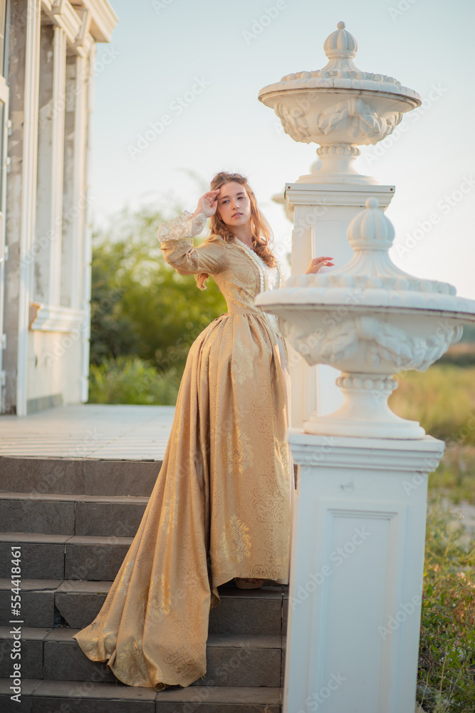 A beautiful young woman in a historic eighteenth century gold dress stands on the stairs of the mansion. Princess in the palace.