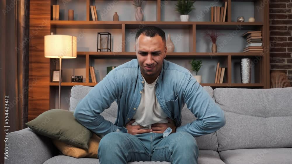 African American man sitting on sofa feeling stomach pain at home, gastritis symptom, peptic ulcer, pancreatitis. Male guy suffering from digestive problems on couch, squeezing belly abdominal pain. African American man sitting on sofa feeling stomach pain at home, gastritis symptom, peptic ulcer, pancreatitis. Male guy suffering from digestive problems on couch, squeezing belly abdominal pain.