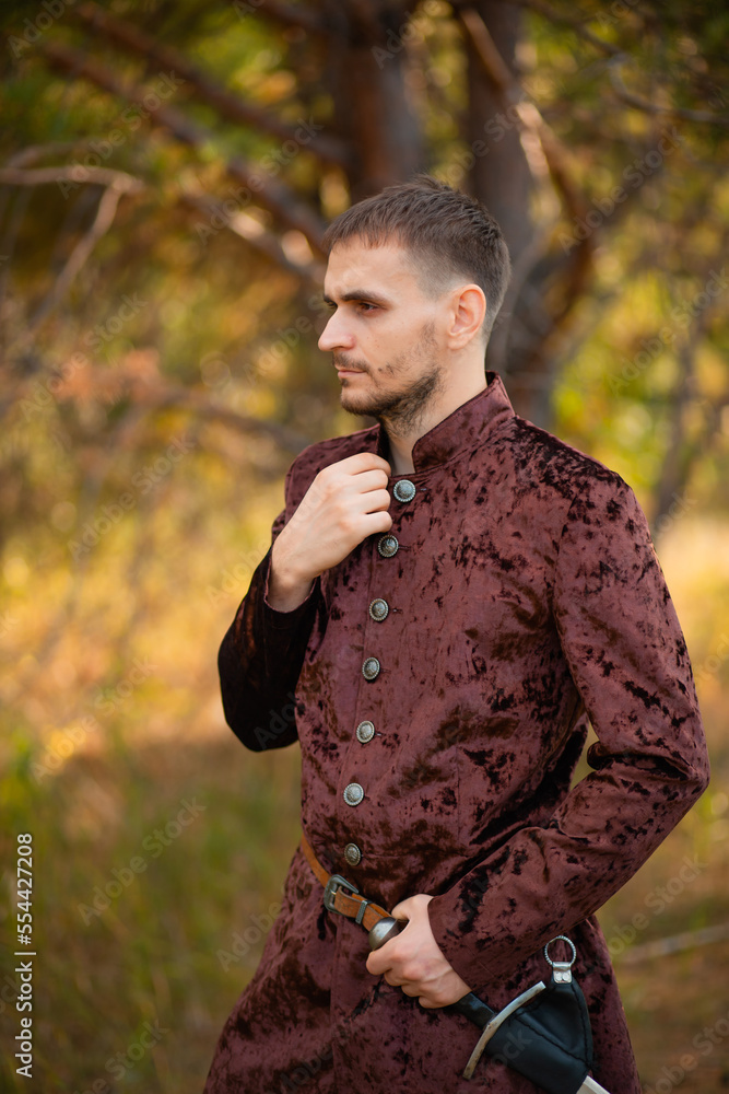 A young man in medieval brown clothes stands in the forest with a dagger. A guy in a historical costume in nature. A young man in medieval brown clothes stands in the forest with a dagger. A guy in a historical costume in nature.