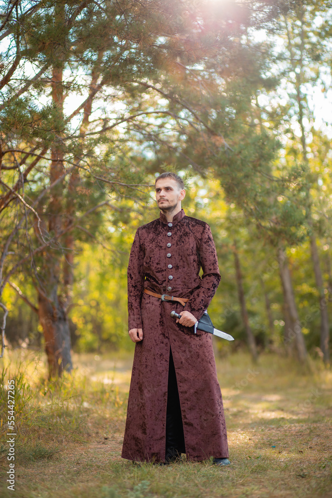 A young man in medieval brown clothes stands in the forest with a dagger. A guy in a historical costume in nature. A young man in medieval brown clothes stands in the forest with a dagger. A guy in a historical costume in nature.