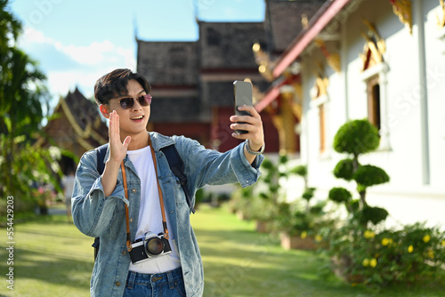 Smiling male travel blogger recording video vlog with smartphone while standing in the buddhist temple