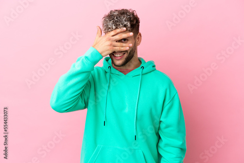 Young handsome caucasian man isolated on pink background covering eyes by hands and smiling