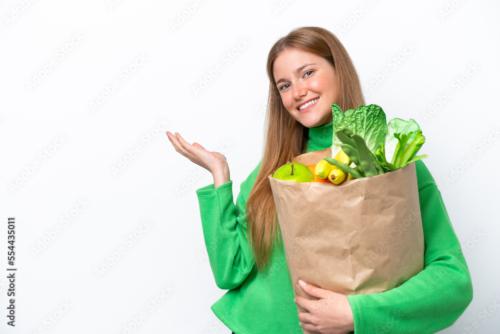 Young caucasian woman holding a grocery shopping bag isolated on white background extending hands to the side for inviting to come Young caucasian woman holding a grocery shopping bag isolated on white background extending hands to the side for inviting to come