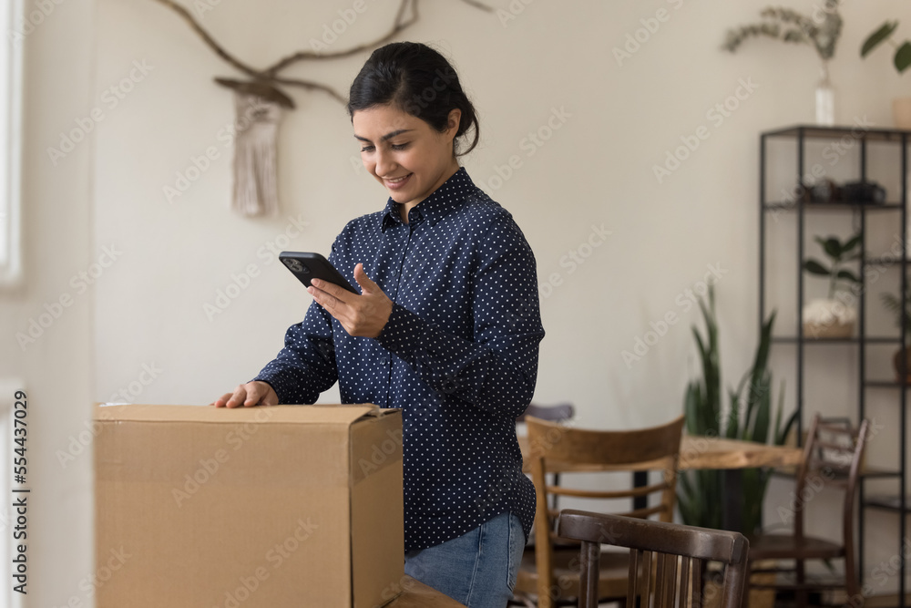 Positive young Indian customer woman packing cardboard box at home, using online on mobile phone, calling for courier, delivery service, unpacking purchase from Internet store Positive young Indian customer woman packing cardboard box at home, using online on mobile phone, calling for courier, delivery service, unpacking purchase from Internet store