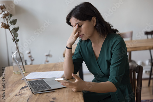 Tired stressed frustrated business woman getting bad news from online chat message, holding smartphone, touching head with closed eyes at work laptop, feeling headache, exhaustion