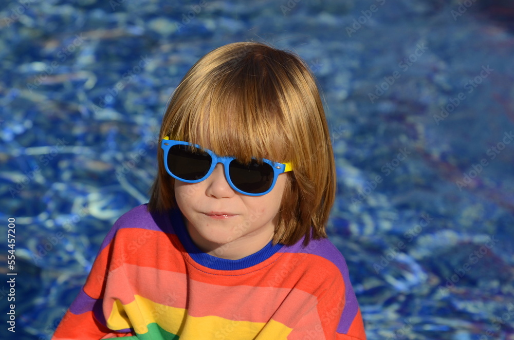 Positive little boy in sunglasses and a multi-colored jacket on the background of the water. Space for text, sun glare on the surface of the pool. Concept: resort, fountain, traveling with children Positive little boy in sunglasses and a multi-colored jacket on the background of the water. Space for text, sun glare on the surface of the pool. Concept: resort, fountain, traveling with children