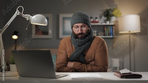 Low air temperature at home. Freezing bearded man in hat and scarf is trying to keep warm. Cold in apartment, in house, indoors