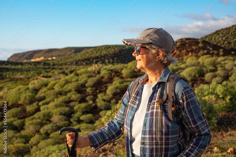 Smiling senior woman in hat and denim shirt walking in outdoor mountain excursion at sunset light enjoying freedom and vacation. Caucasian female having healthy lifestyle in retirement