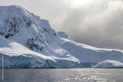 Soaring peak in the Antarctic Peninsula, sloping towards the sea. Rocks visible through the pristine snow cover. Calm sea reflecting sunlight in the foreground. Clouds with sunbeams in the background. © dhayes