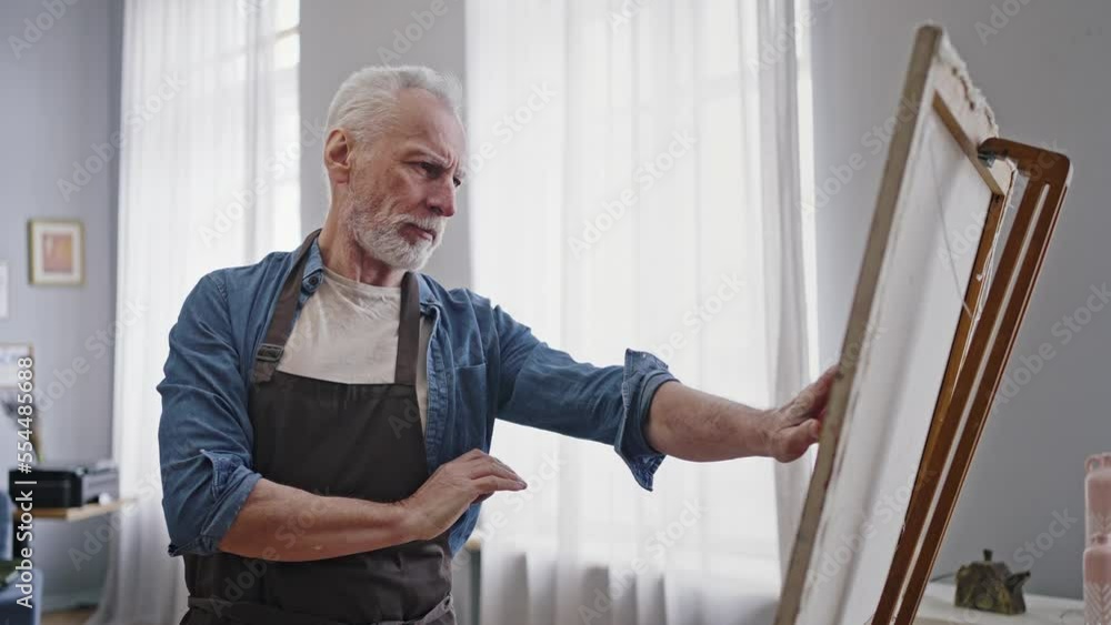Elderly artist with crossed arms, looking at his artwork, taking out a canvas from a wooden easel Elderly artist with crossed arms, looking at his artwork, taking out a canvas from a wooden easel