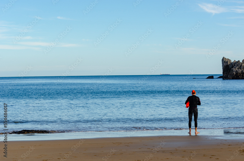 Persona con neopreno negro, gorro rojo y salvavidas rojo preparado para nadar en el mar