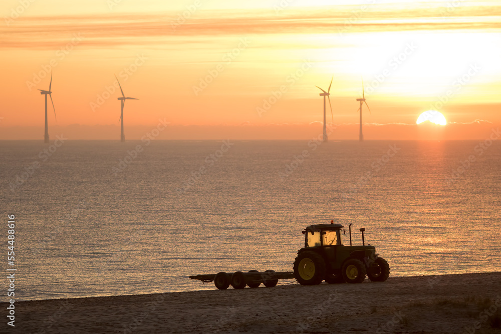 Fototapeta premium Misty morning sunrise. Beach tractor with offshore wind farm turbines. Eco landscape image for environmental conservation.