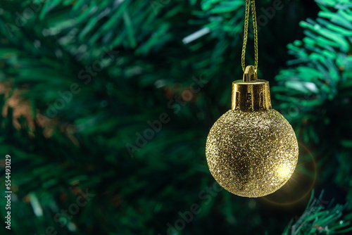 Uma bola dourada, pendurada no galho com detalhes da árvore de natal ao fundo.