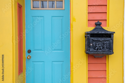 The exterior wall of colorful orange and yellow wooden clapboard siding building with a bright blue door.   A black decorative metal postal or letterbox is affixed to the house with a lock mechanism.