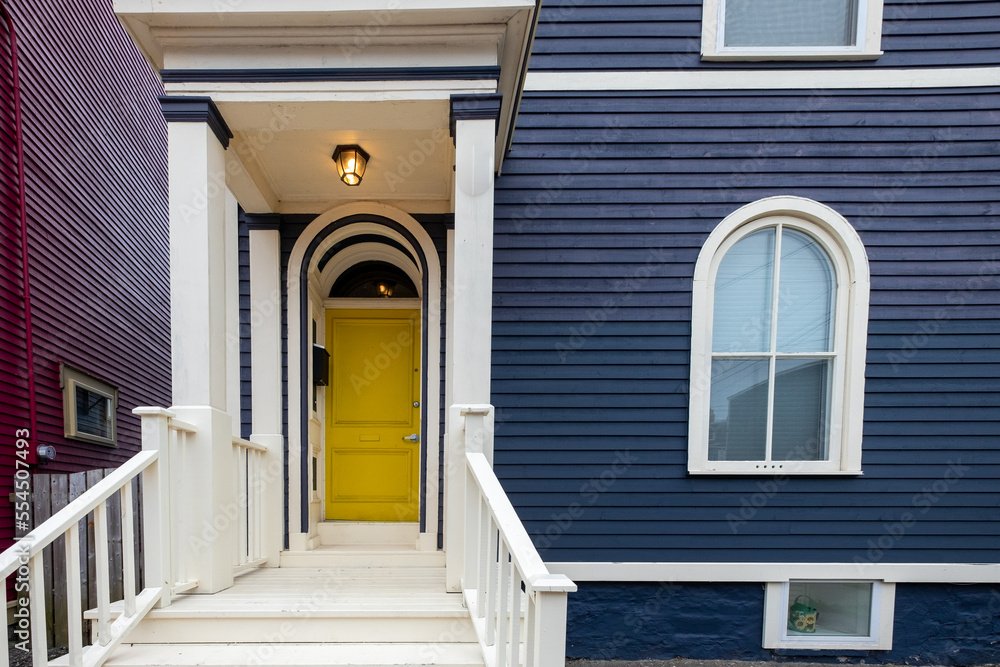 A rounded top bright yellow elegant style door in a dark blue colored wooden house. The residence has a four pane half-moon glass window with cream color trim. The stairs at the entrance are wooden.