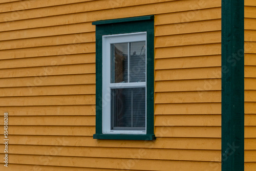 The exterior corner wall of a vibrant yellow colored wood clapboard siding. There's a single double hung window with green and white trim in the house. The corner of the building is thick green board.