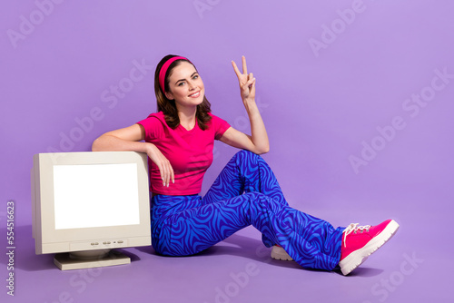 Photo of cheerful cute lady sitting floor demonstrates old computer empty space novelty v-sign isolated on purple color background