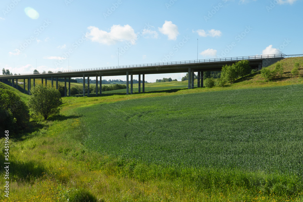 Eine Brücke über ein kleines Tal in einer ländlich geprägten Landschaft mit Wiese, jungen Getreide, Bachlauf in der Nähe von Jessheim, Norwegen.