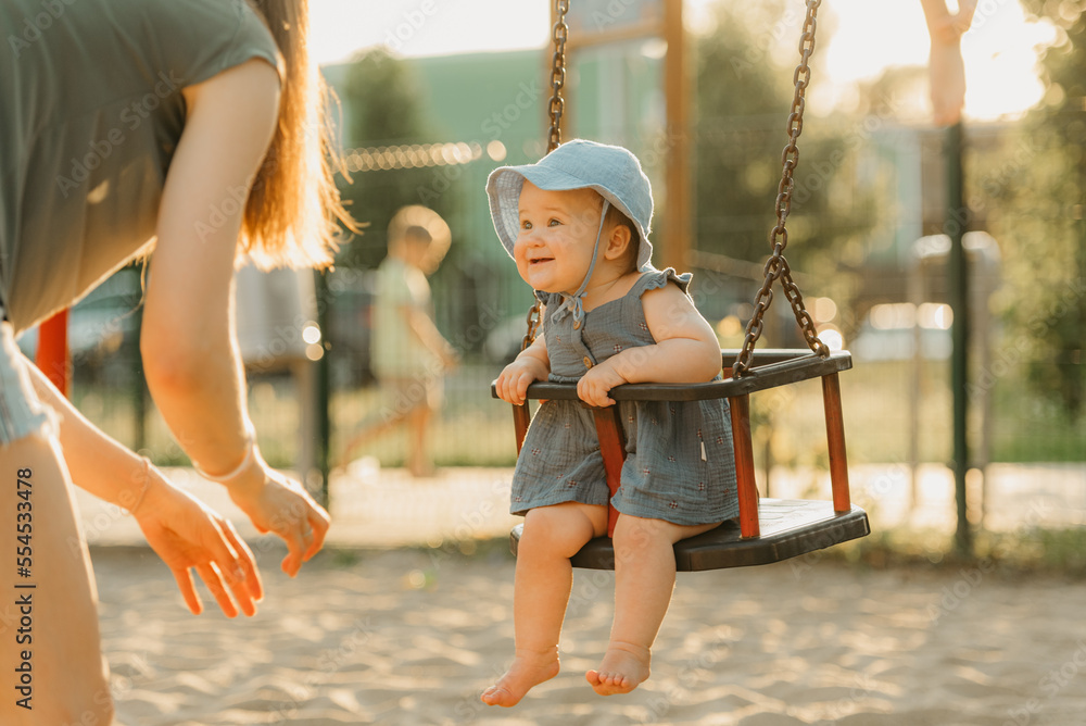 Toddler baby girl in a dress is joying on a swing on the warm summer evening