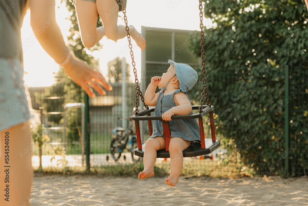 Baby girl in a dress is sucking her thumb on a swing on the warm summer evening