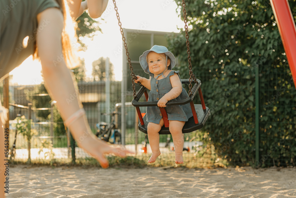 Toddler baby girl in a dress is laughing on a swing on the warm summer evening