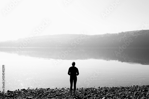 Woman gazes across the still morning water of the Puget Sound in Washington State with the evergreen covered hills reflecting off the clear water.