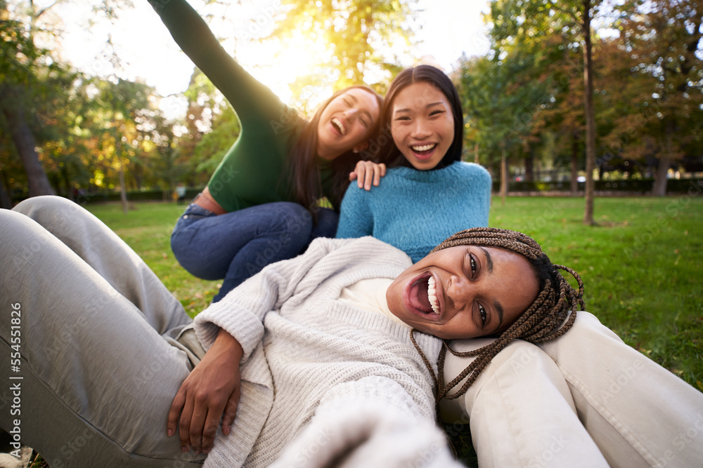 Naklejka premium Three multiracial girls lying on the grass taking a selfie looking at the camera. Group of cheerful friends having a fun together, sharing happiness on social media. Concept of friendship and free