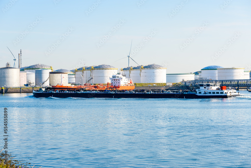 Tanker barge sailing past an oil dock with large steel fuel tanks on a clear summer day