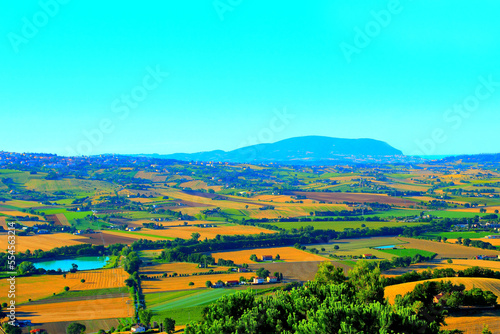 Serene scene in Montelupone with the typical Marche hilly landscape and various fields filling the picture, a body of water, a densely populated human settlement, and Monte Conero in the background photo