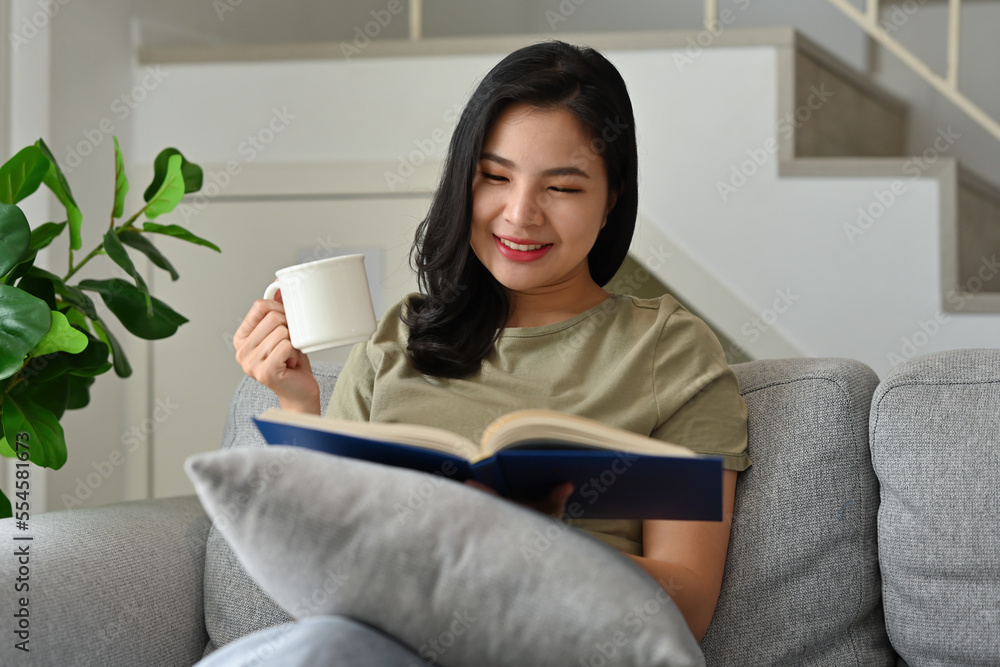 Millennial Asian girl sitting on sofa enjoy with morning coffee while reading a book at home. Millennial Asian girl sitting on sofa enjoy with morning coffee while reading a book at home.