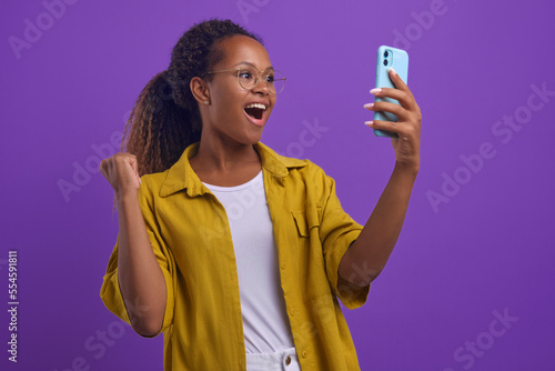 Young overjoyed attractive African American woman holds mobile phone and makes victorious wave of hand after receiving message with date invitation in social network stands on lilac background
