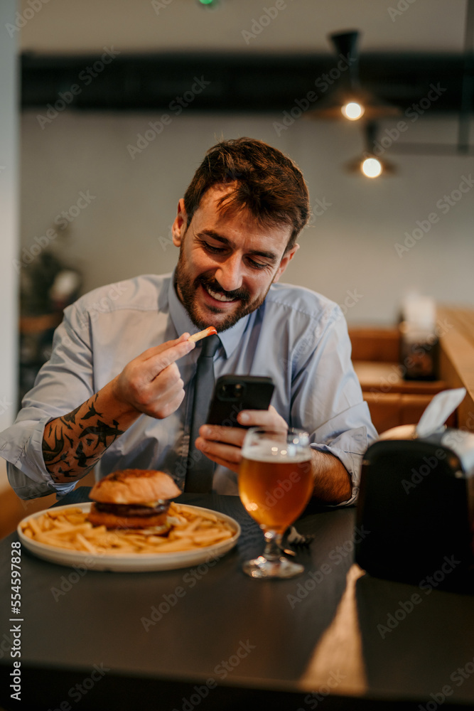 Focused businessman sitting in the local cafe, having a lunch break with a burger and beer and texting on his phone. Focused businessman sitting in the local cafe, having a lunch break with a burger and beer and texting on his phone.