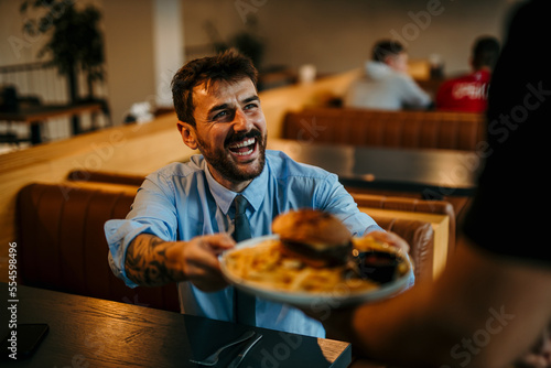 The waiter is serving a delicious burger and french fries to a smiling businessman who is having lunch alone.