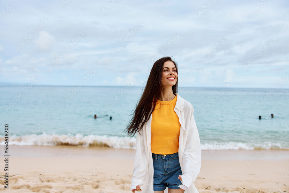Portrait of a happy woman smile with teeth with long hair brunette walks along the beach in a yellow tank top denim shorts and a white shirt by the sea summer travel and feeling of freedom, balance