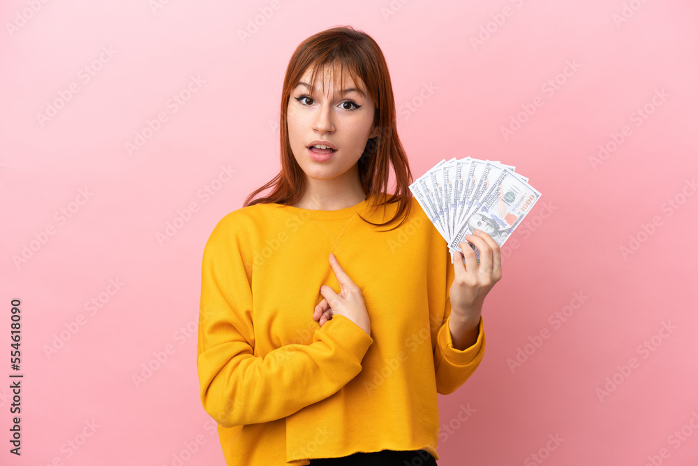 Redhead girl taking a lot of money isolated on pink background with surprise facial expression Redhead girl taking a lot of money isolated on pink background with surprise facial expression