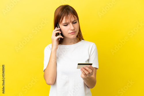 Redhead girl isolated on yellow background buying with the mobile with a credit card