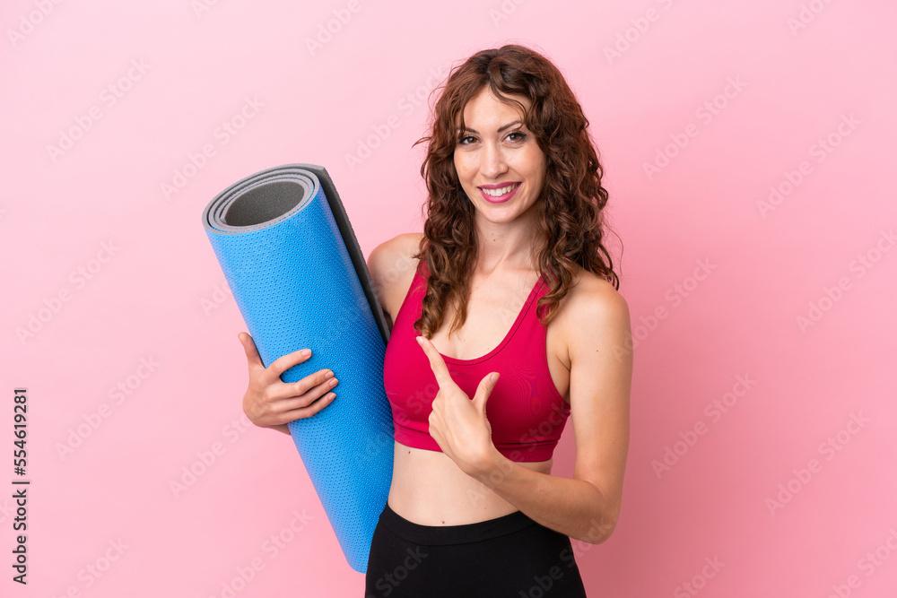 Young sport woman going to yoga classes while holding a mat isolated on pink background pointing back Young sport woman going to yoga classes while holding a mat isolated on pink background pointing back