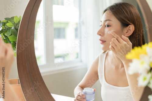 Fresh skin, beautiful smile of asian young woman, girl looking at mirror, hand applying moisturizer lotion on her face, putting cream treatment before makeup cosmetic routine at home. Facial Beauty.
