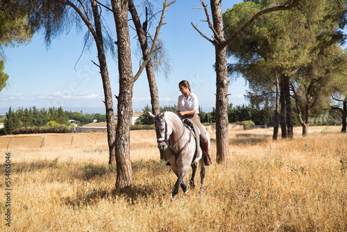 Beautiful young woman walking, riding her horse in the countryside next to several pine trees, on a sunny day. Concept horse riding, animals, dressage, horsewoman. © Manuel