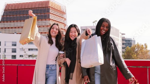 Three multiethnic young women smiling showing their new shopping bags. Group of happy shopaholic girls excited in the sale week. Consumism Concept. High quality 4k slow motion footage photo