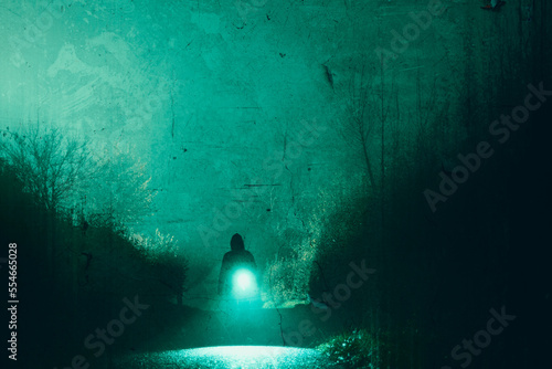 A scary hooded figure carrying a torch, silhouetted along a mysterious country road. On a spooky foggy winters night. With a grunge edit. © Dave