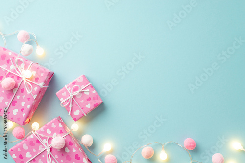 Valentine's Day concept. Top view photo of gift boxes in wrapping paper with heart pattern light bulb garland and soft pompons on isolated pastel blue background with copyspace © ActionGP