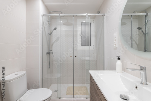 Spacious white ceramic tile bathroom is illuminated by bright white natural light. Large round mirror on the wall reflects the shower area with glass railing and metal fittings.