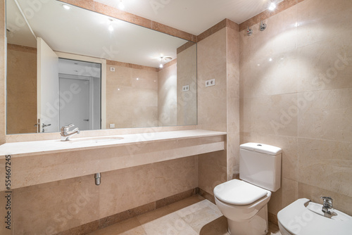 Modern spacious bathroom with bright beige tiles  toilet  bidet and sink on long marble countertop. Full-length mirror reflects open door to hallway. Bright light from bulbs on ceiling.