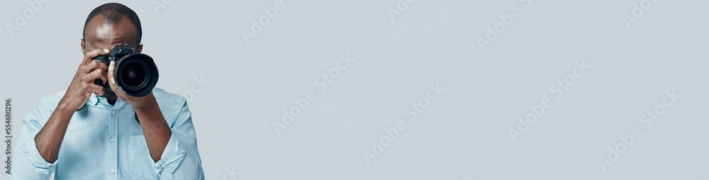 Young African man taking a photo using digital camera while standing against grey background Young African man taking a photo using digital camera while standing against grey background