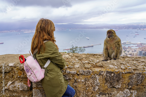 Female tourist on her back watching a small Gibraltar monkey high on the rock with the bay in the background.