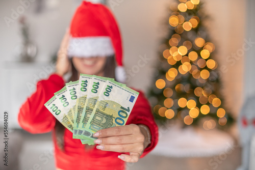 Frau mit Weihnachtsmütze und Geld als Geschenk in der Hand. Bokeh von den Lichtern eines Weihnachtsbaums im Hintergrund.