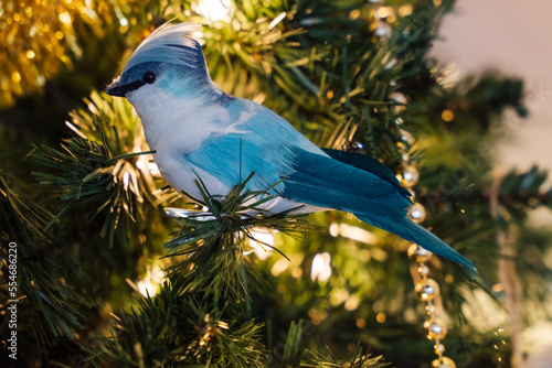 Blue Jay Christmas ornament perched in an artificial Christmas tree with Christmas light in background