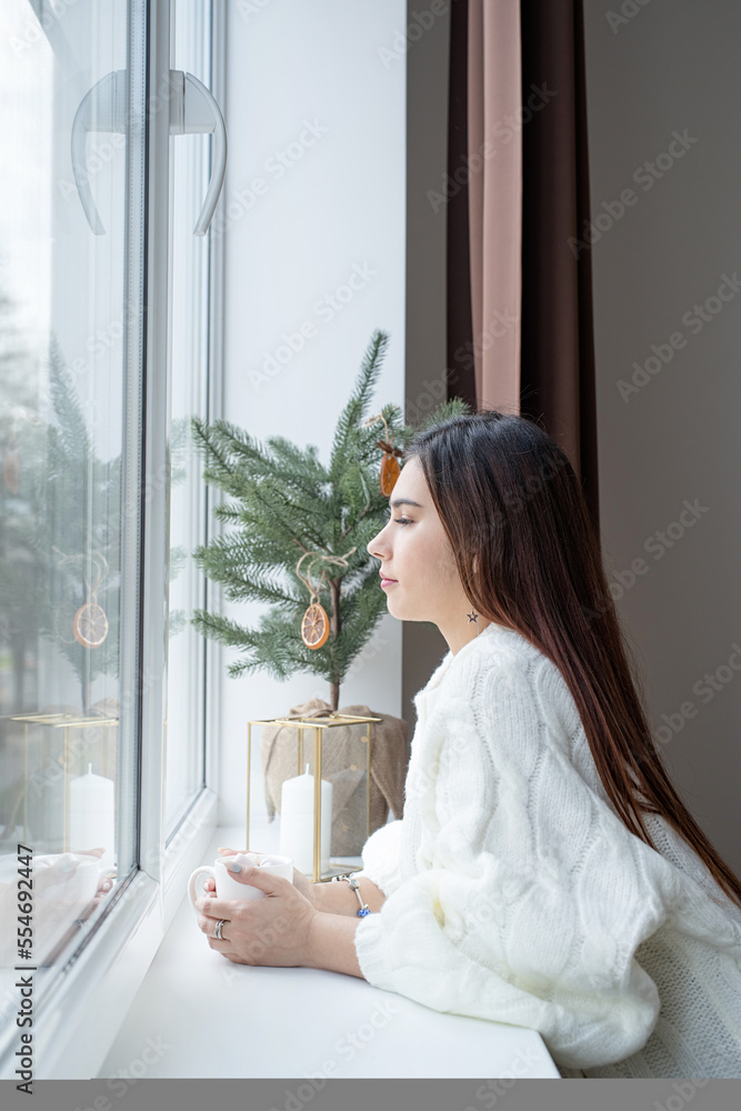 Woman in warm white winter sweater standing next to the window at home at christmas eve holding cup with marshmallows, fir tree behind
