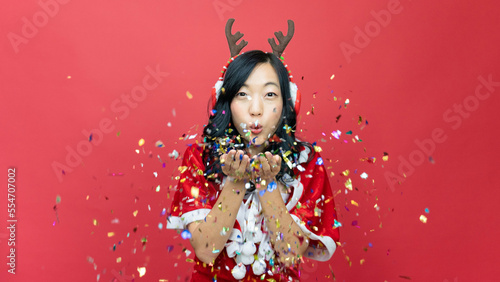 Happy lovely woman with red santy costume sending a kiss to camera on red background with confetti. Concept of the New Year and Christmas Day.